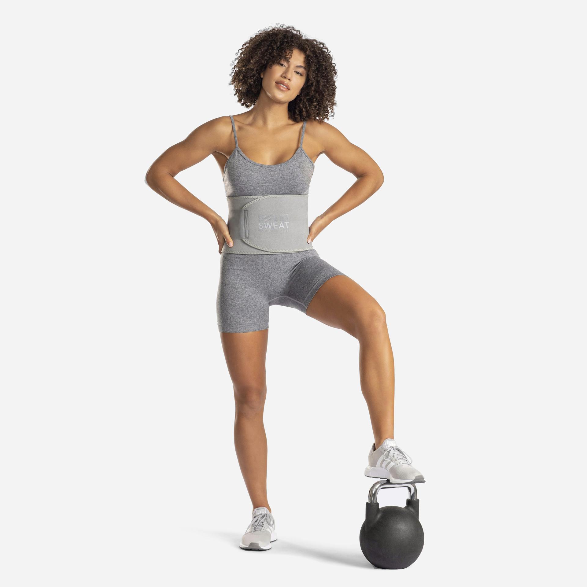 Woman in workout attire resting a foot on a weighted kettlebell, while wearing a Sweet Sweat® Matte Series waist trimmer.